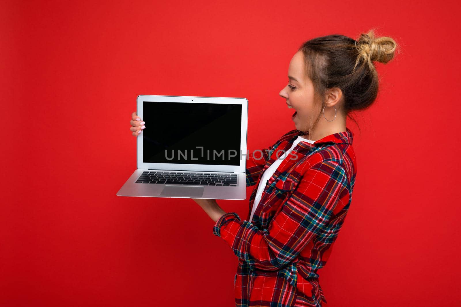 side profile photo of Beautiful amazed young woman holding laptop wearing red shirt looking at monitor display isolated on red background. Mock up