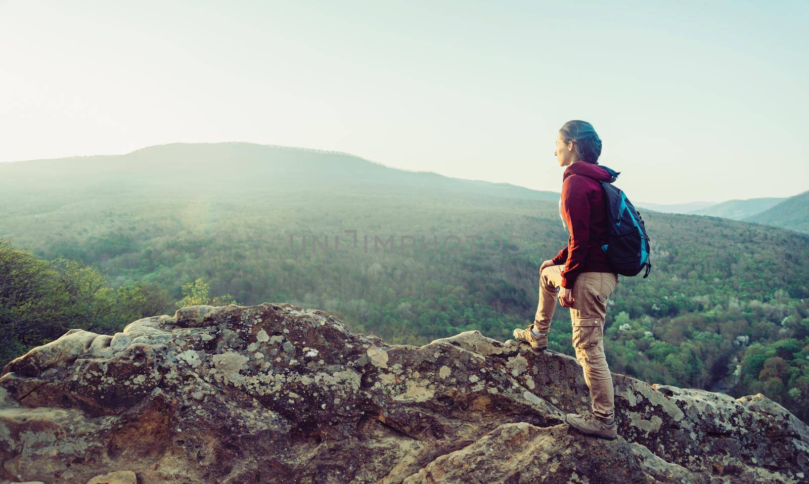 Hiker young woman with backpack standing on peak of rock in the mountains and looking into the distance in summer outdoor