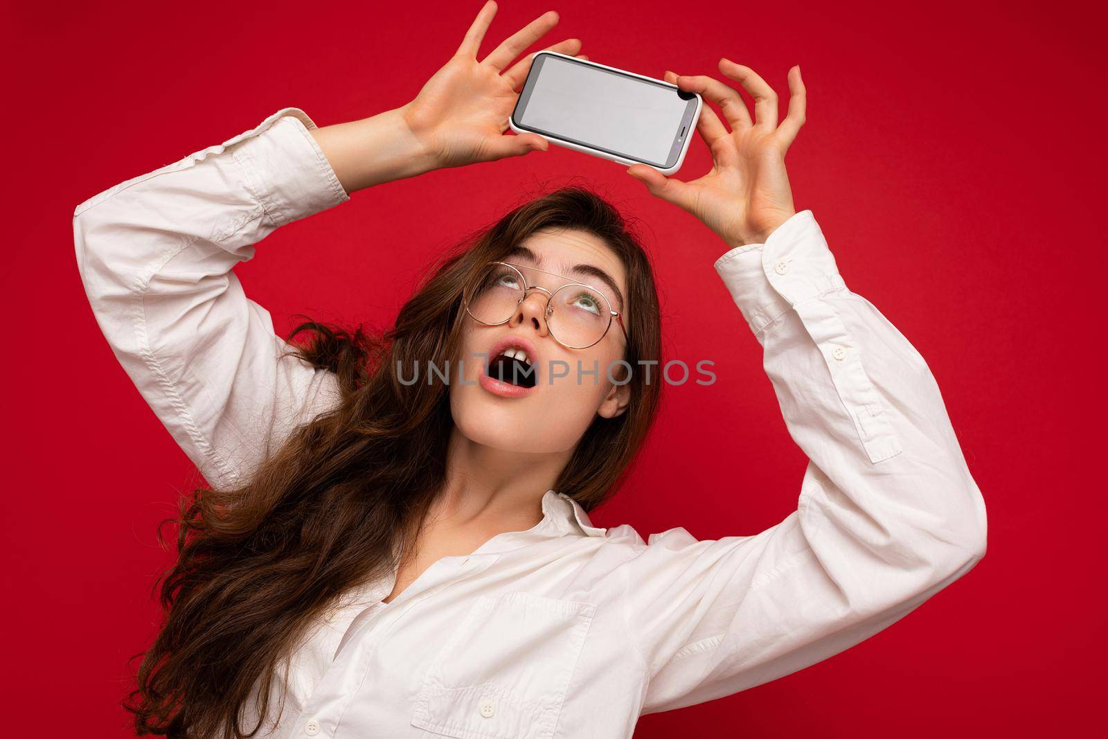 attractive amazed young brunet woman wearing white shirt and optical glasses isolated over red background holding in hand and showing mobile phone with empty screen for mockup looking at gadjet screen.