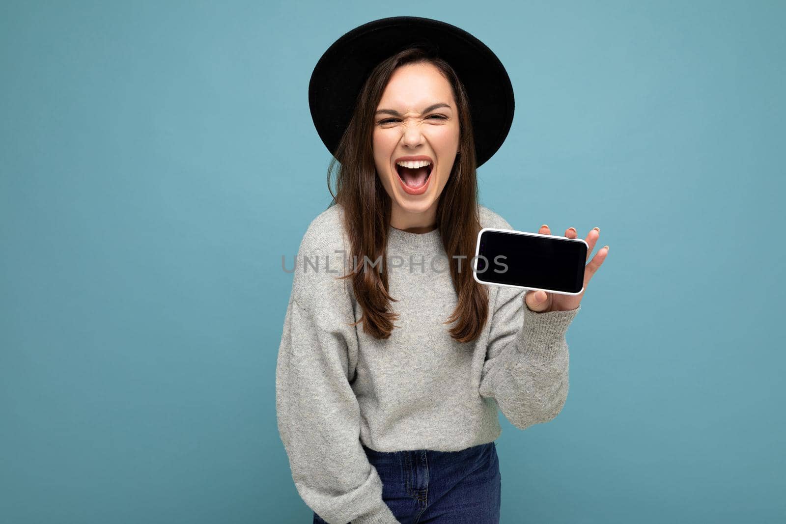 Portrait photo shot of beautiful funny young woman wearing black hat and grey sweater holding phone showing smartphone isolated on background.Mock up, cutout, copy space
