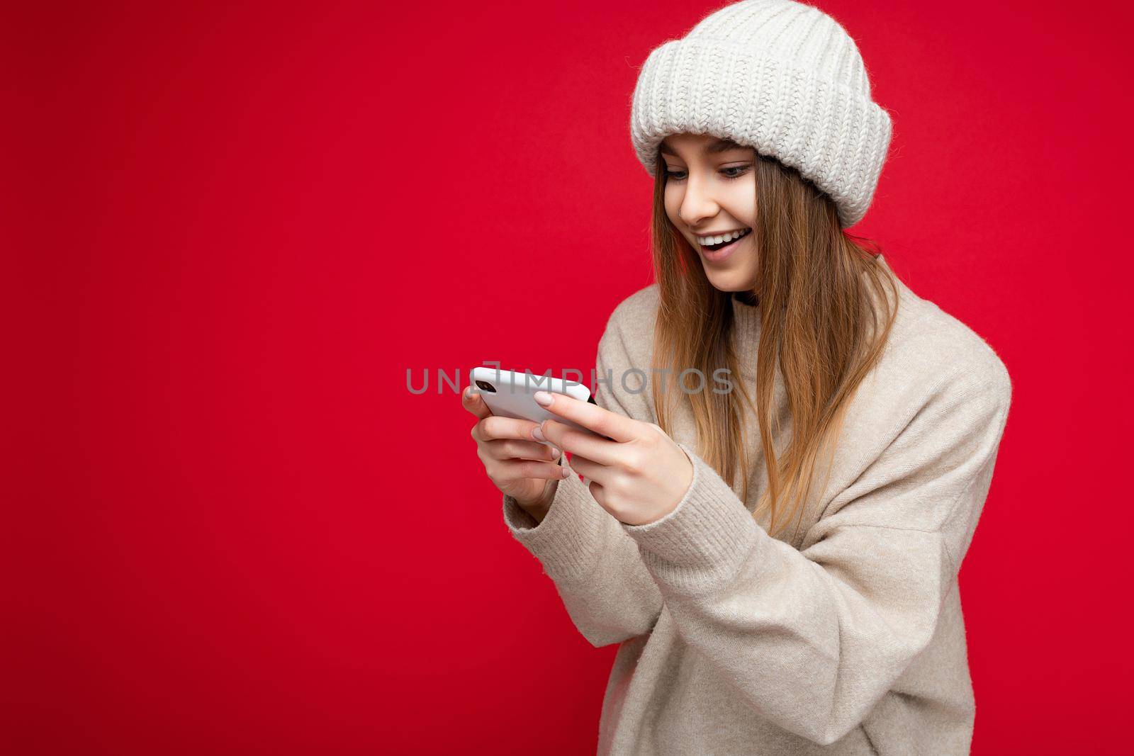 Beautiful young blonde woman wearing casual beige sweater and beige hat isolated over red background holding in hand and using mobile phone communicating online on the internet looking at gadjet display. copy spca
