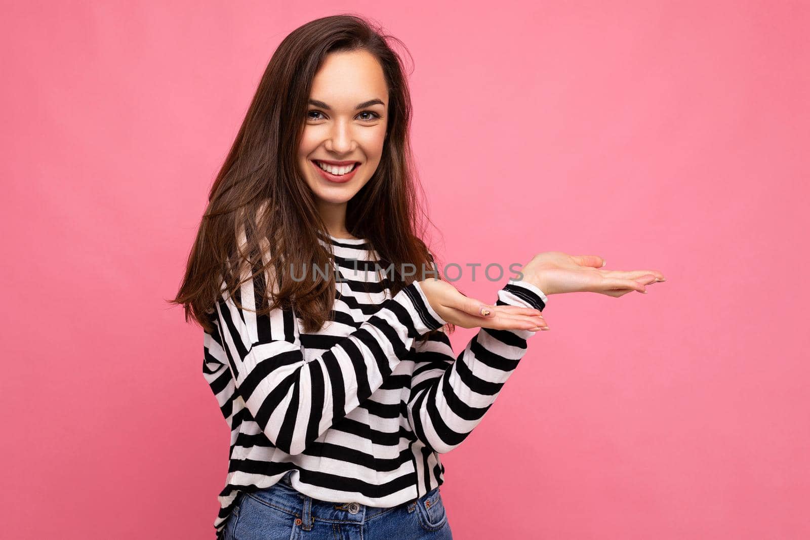 Photo of young happy positive beautiful brunette woman with sincere emotions wearing casual striped pullover isolated on pink background with copy space and pointing at empty space for advertisement and news.