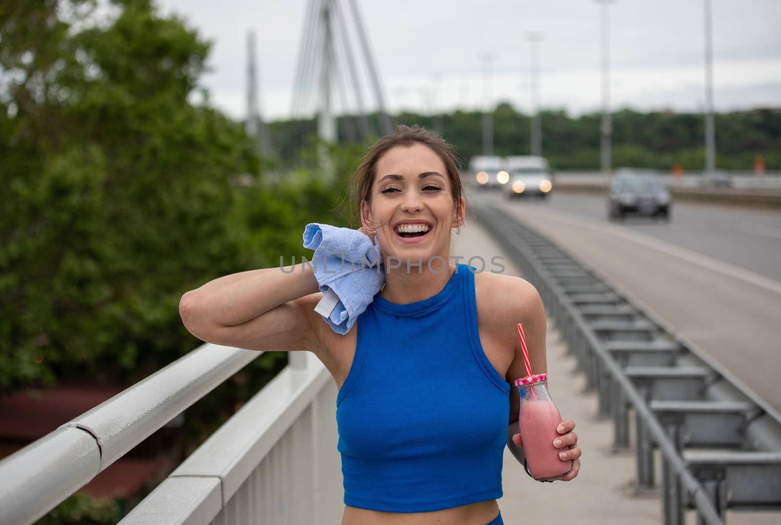 Young woman resting after workout wiping sweat smiling. Athlete hydrating having strawberry milkshake on bridge.