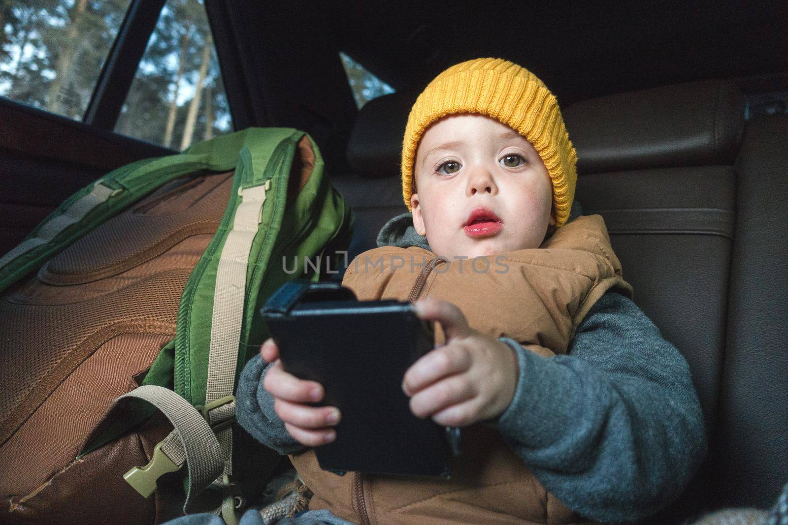 Charming toddler in outwear and hat sitting on backseat in car with device in hands looking at camera.