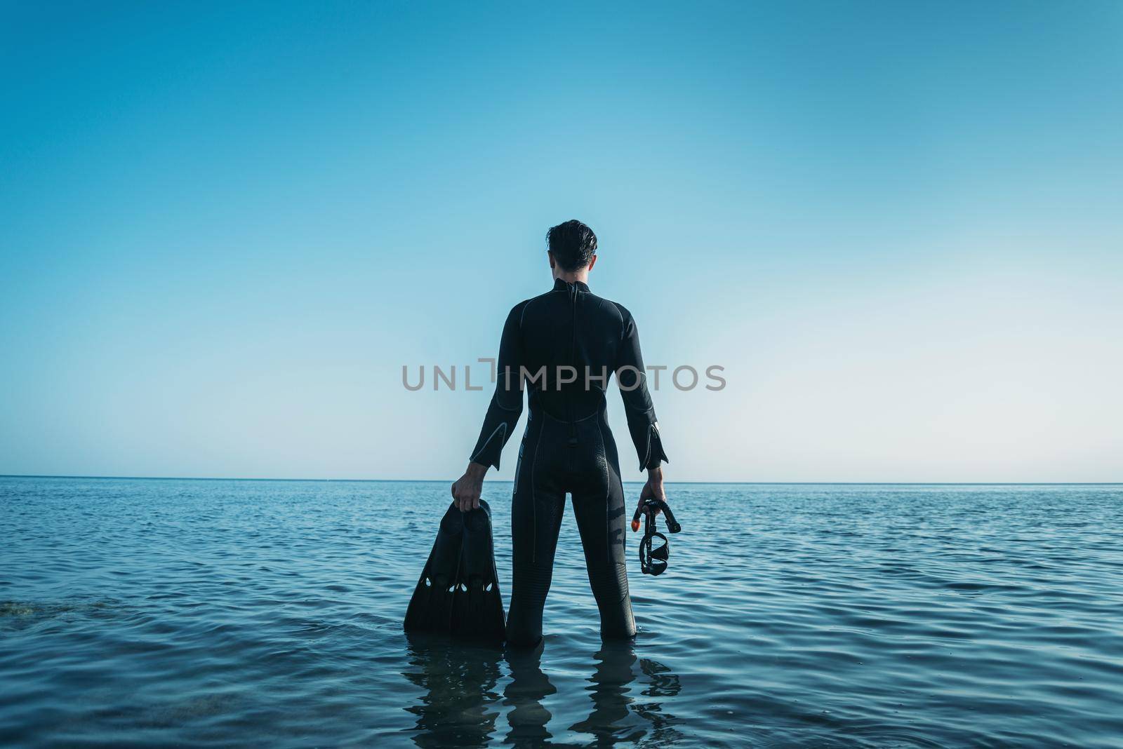 Young man wearing in a diving suit with a mask and flippers standing in the sea and looking into the distance in summer, rear view