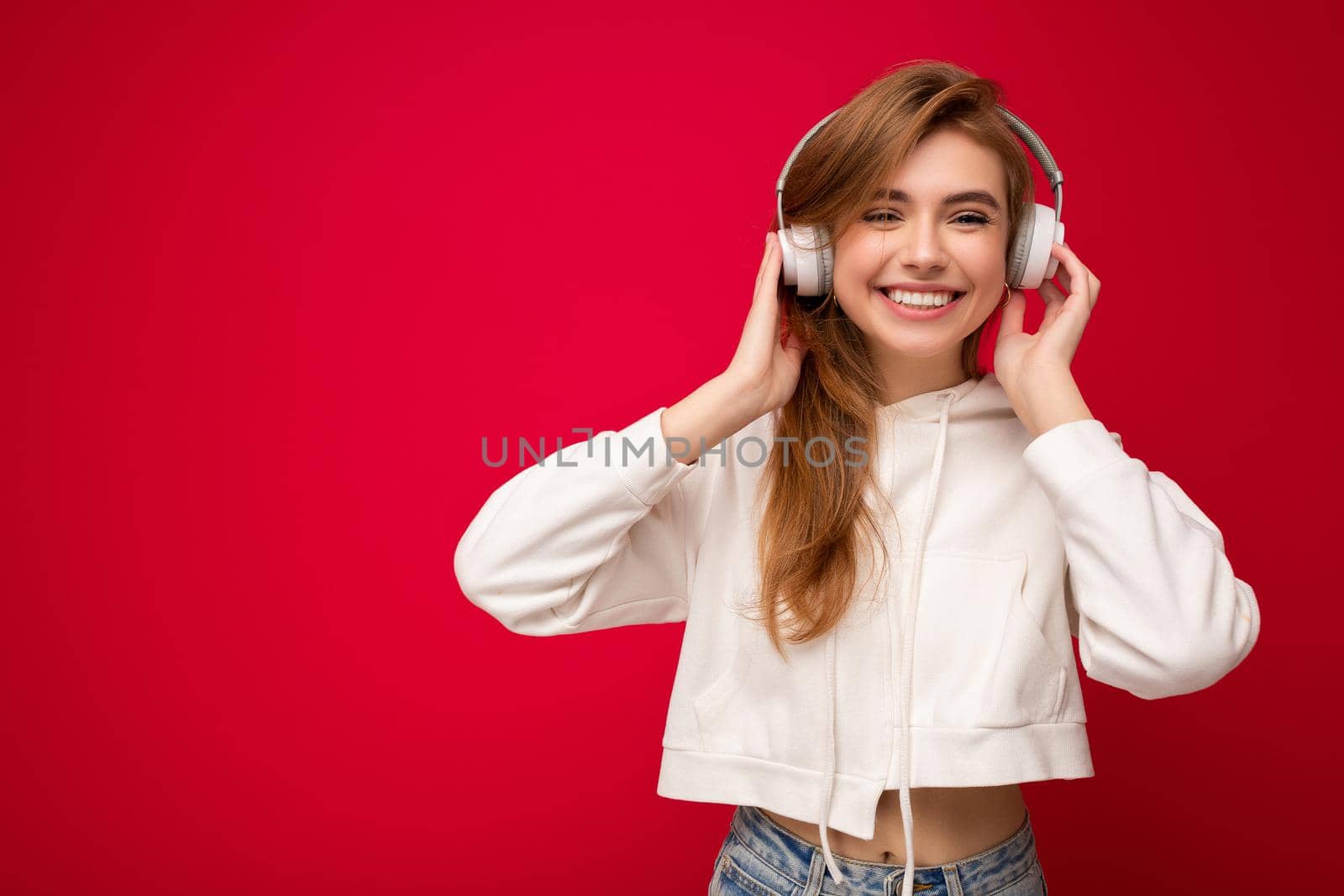 Photo of beautiful happy smiling young blonde woman wearing white hoodie isolated over colourful background wall wearing white wireless bluetooth earphones listening to cool music and enjoying looking at camera. cope space