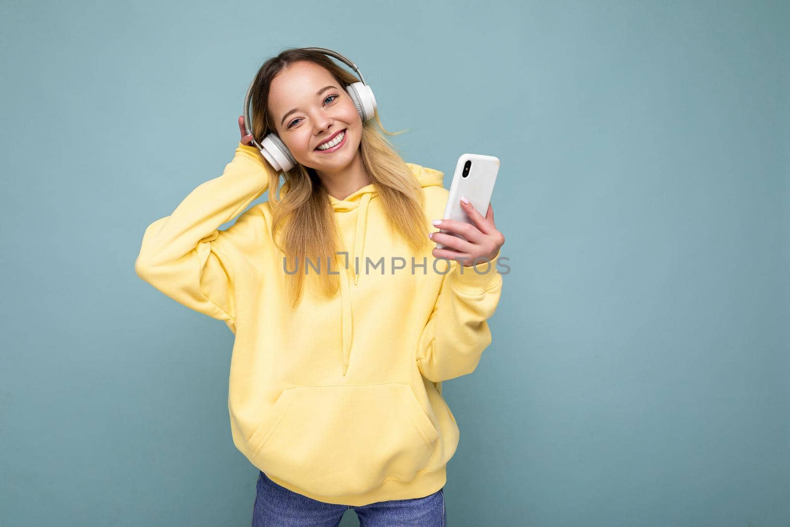 Photo shot of beautiful joyful smiling young female person wearing stylish casual outfit isolated over colorful background wall wearing white bluetooth wireless earphones and listening to music and using mobile phone looking at camera.