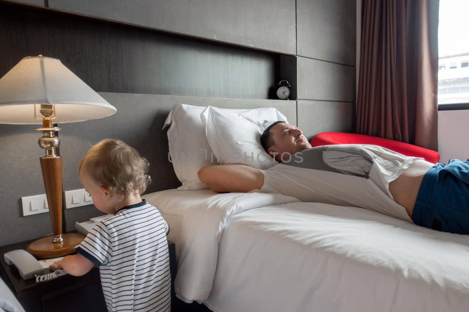 Content father lying on cozy bed in hotel room with cute infant playing near.