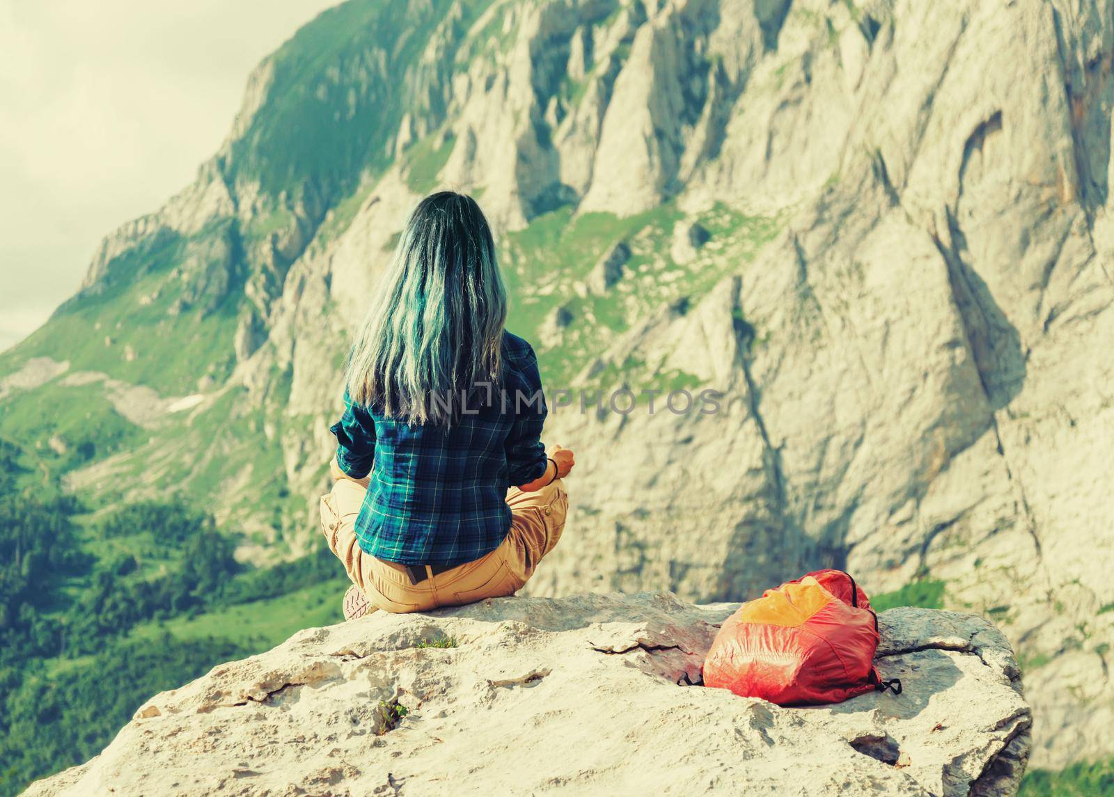 Traveler young woman sitting on rock stone in summer mountains, rear view. Image with instagram color