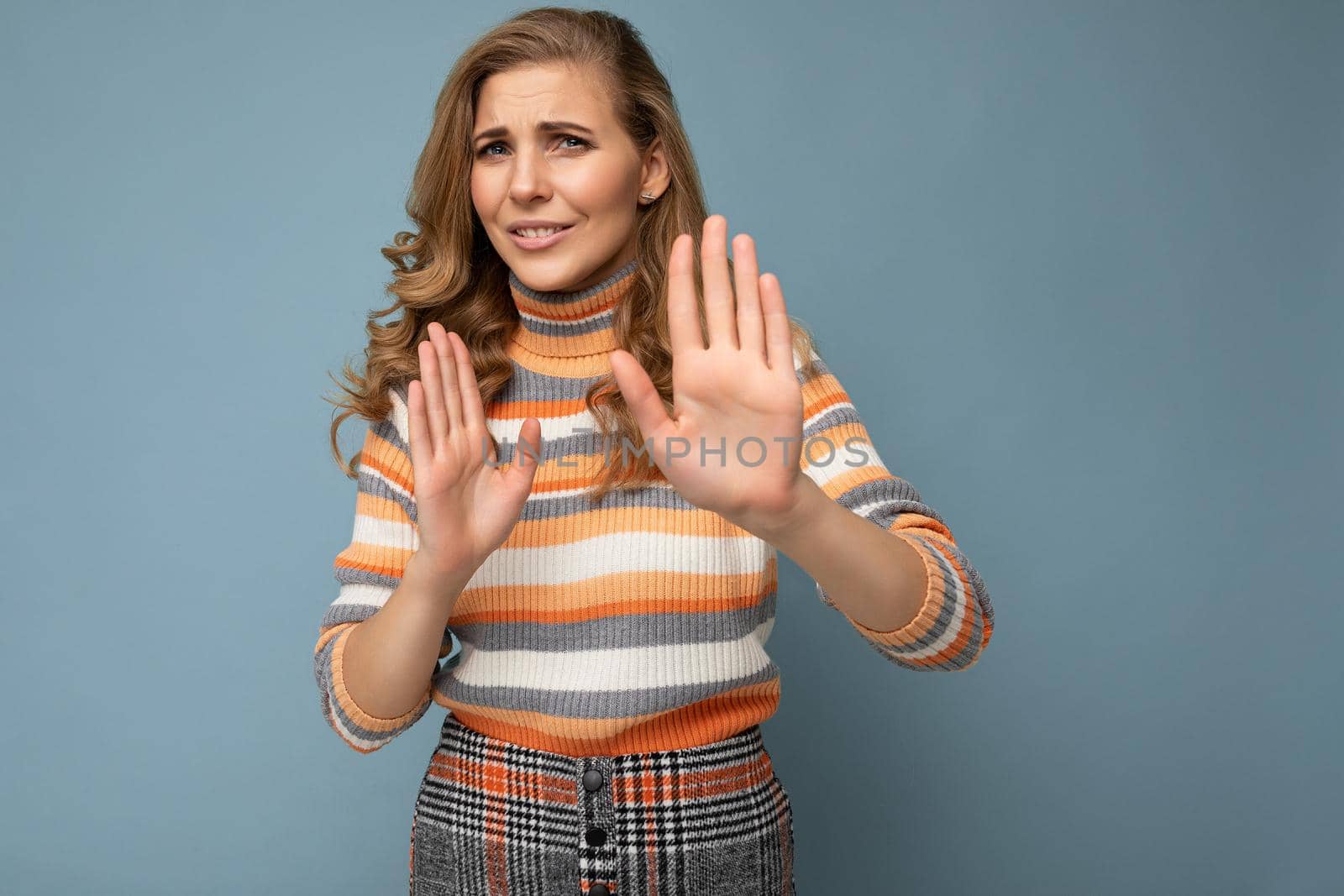 Portrait of young pretty beautiful sad angry dissatisfied blonde woman with sincere emotions wearing casual striped sweater isolated over blue background with copy space and showing no gesture and stop sign with palms. Negative concept.