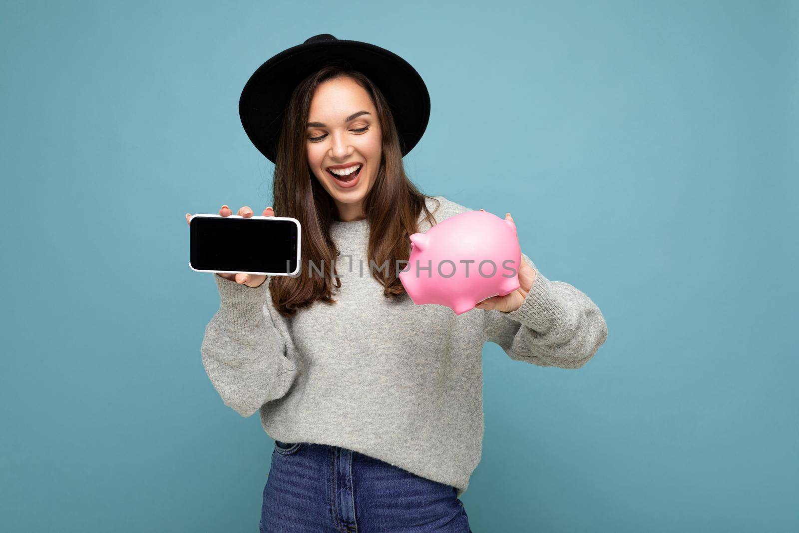 Portrait of happy positive smiling young beautiful brunette woman with sincere emotions wearing stylish gray pullover and black hat isolated on blue background with copy space and holding pink pig penny bank and showing mobile phone with empty screen display.