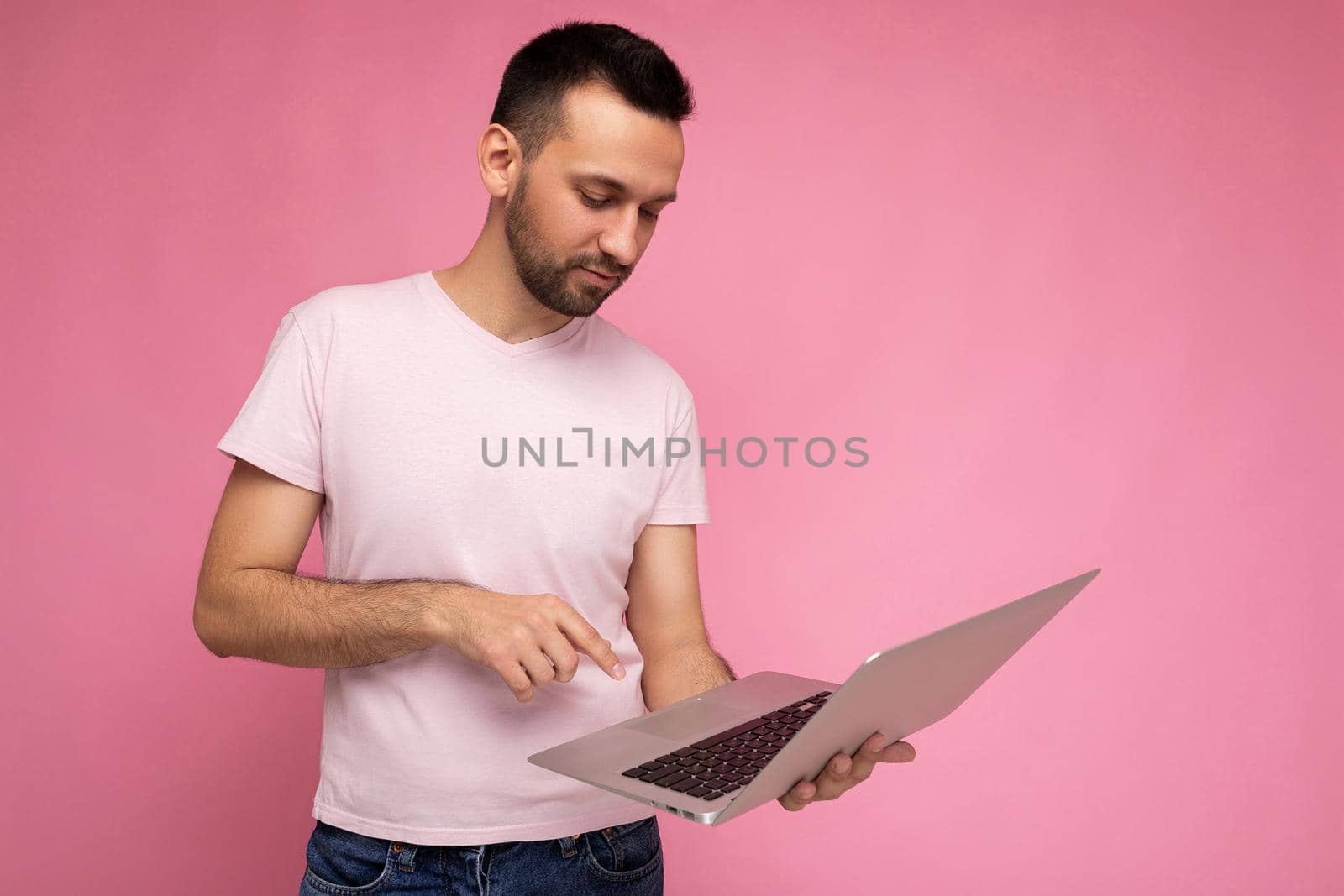 Handsome brunet man holding laptop computer and typing on the keyboard looking at netbook monitor in t-shirt on isolated pink background.