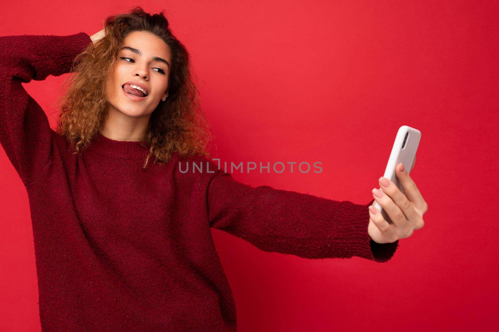 Closeup of Sexy charming Beautiful young woman with curly hair wearing dark red sweater isolated on red background wall holding and using smart phone looking at telephone screen and taking selfie.