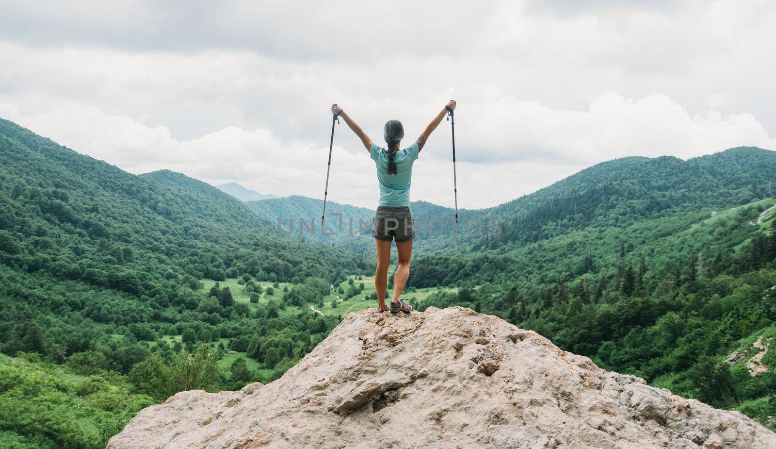 Happy hiker young woman with trekking poles standing on peak of cliff in summer mountains