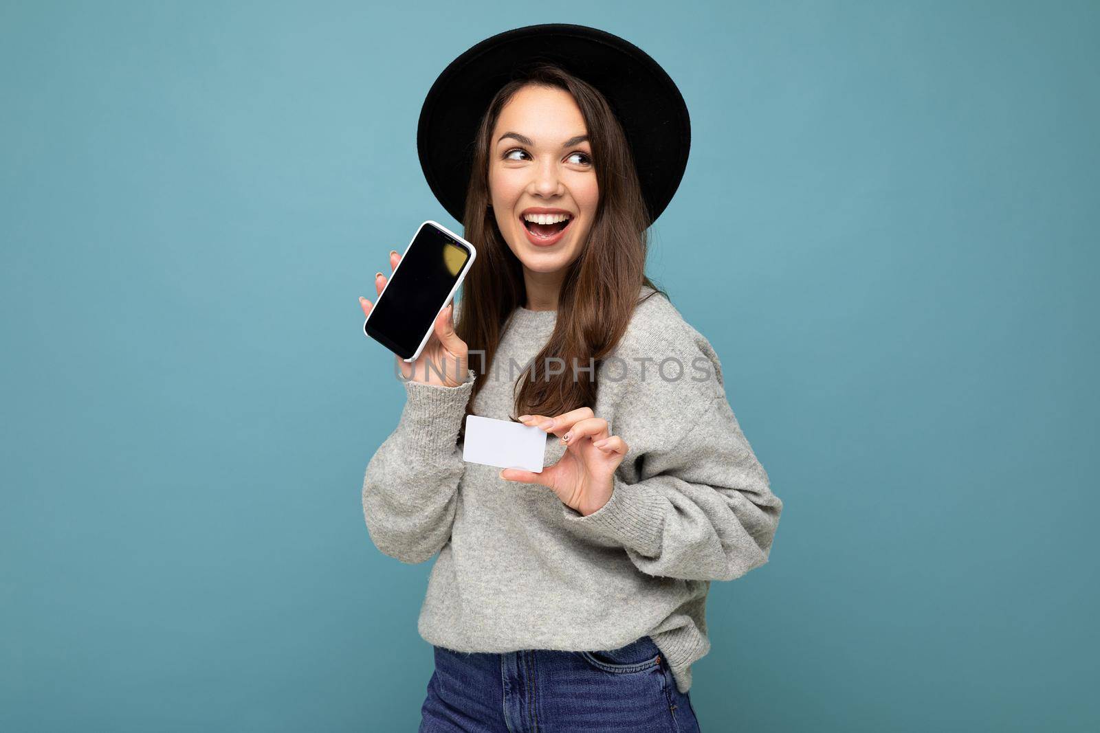 Photo of attractive joyful young brunette woman wearing black hat and grey sweater isolated over blue background holding credit card and mobile phone with empty display for mockup looking to the side.