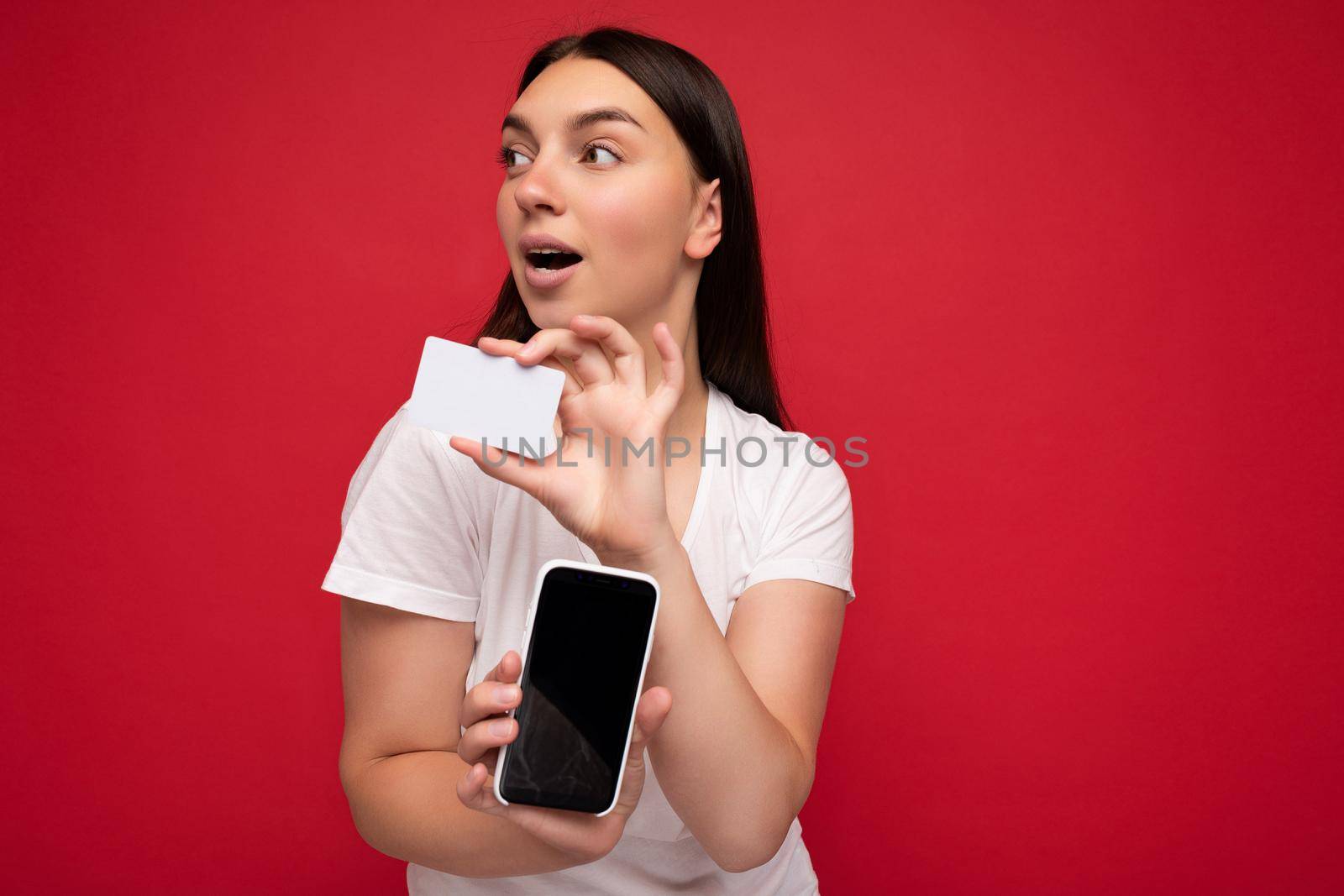 Photo of attractive happy young brunette woman wearing casual white t-shirt isolated over red background with empty space holding in hand mobile phone and showing smartphone with empty screen for mockup and credit card looking to the side.