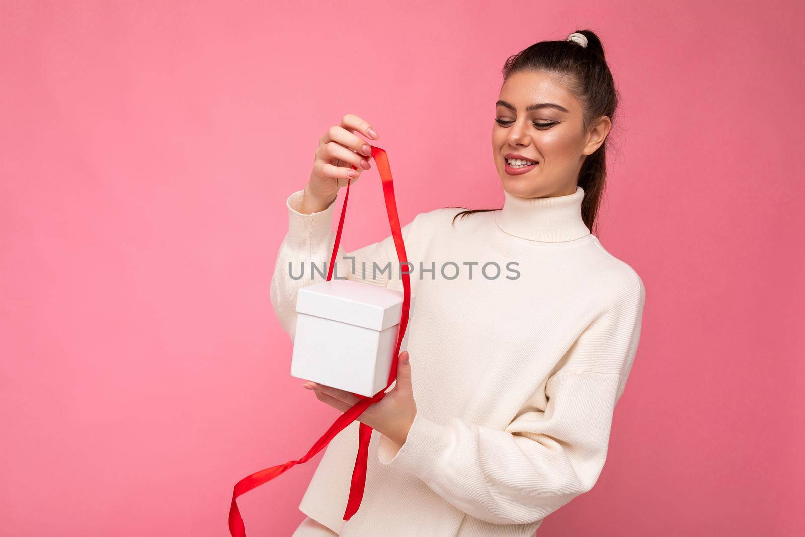 Beautiful happy young brunette woman isolated over colourful background wall wearing stylish casual clothes holding gift box and looking to the side.