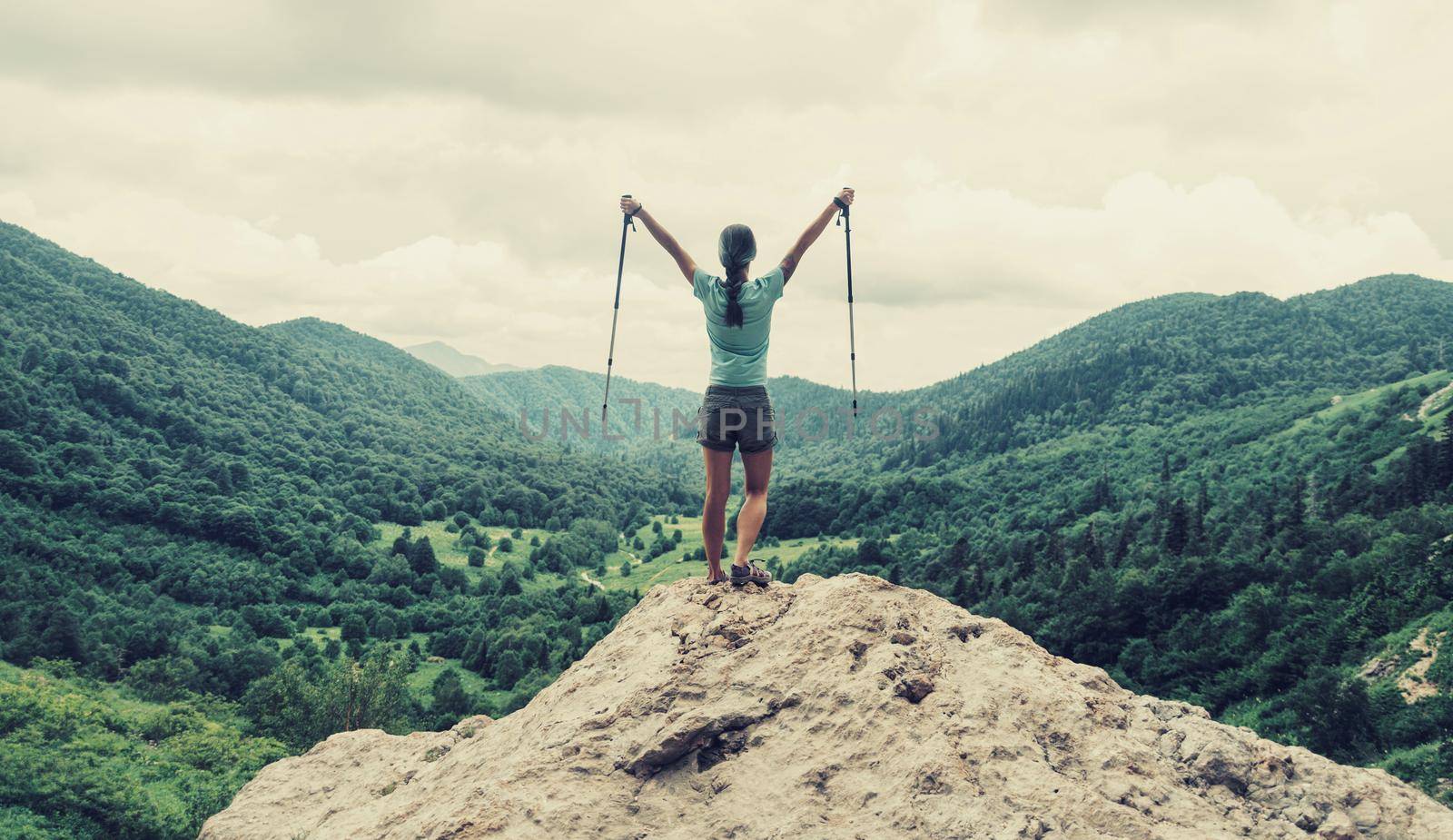 Happy traveler young woman with trekking poles standing on peak of cliff in summer mountains