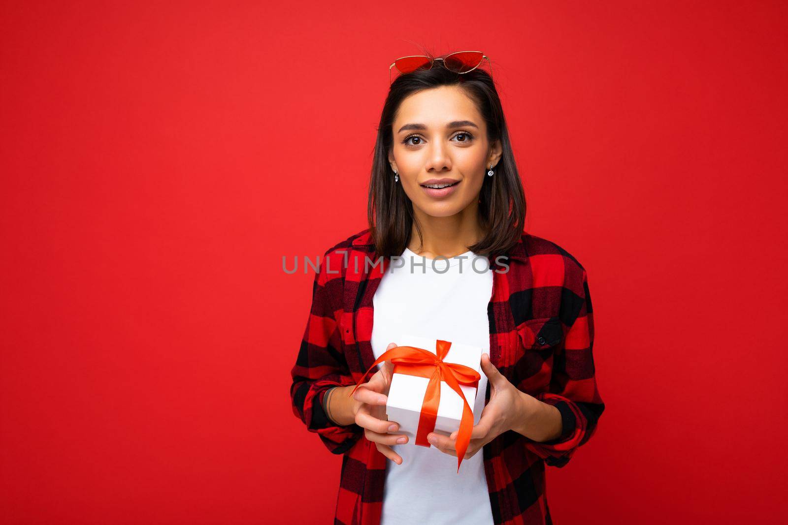 Photo shot of pretty positive young brunette woman isolated over red background wall wearing white casual t-shirt and red and black shirt holding white gift box with red ribbon and looking at camera.