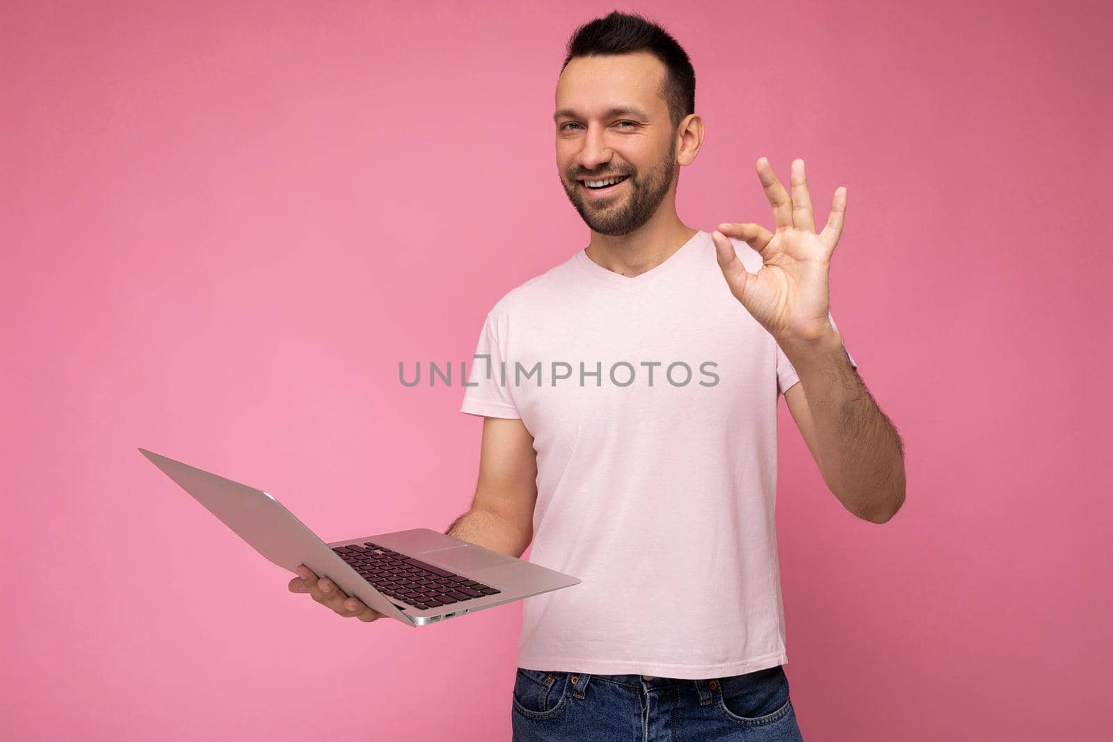 Handsome happy and funny brunet man holding laptop computer and showing gesture okay looking at camera in t-shirt on isolated pink background.