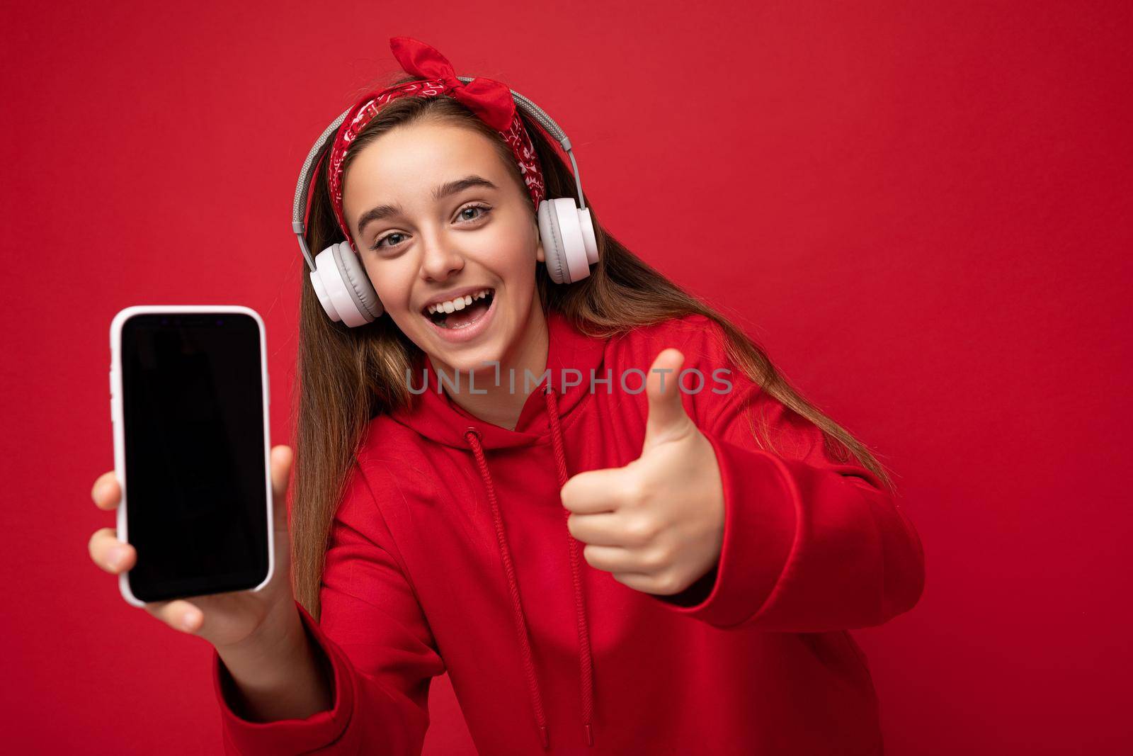 Attractive joyful smiling brunette girl wearing red hoodie isolated on red background holding and showing smartphone with empty display for cutout wearing white wireless headphones listening to funny music looking at camera and showing thumbs up gesture.