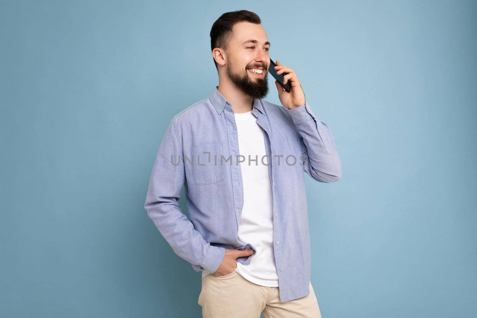 Side-profile Photo shot of good looking young brunet bearded man wearing casual blue shirt and white t-shirt poising isolated on blue background with empty space holding in hand and communicating talking on mobile phone looking to the side.