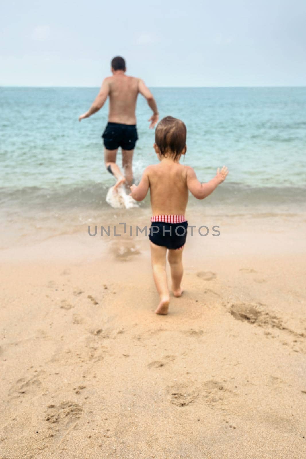 Back view of little child and father running together on wet sandy beach