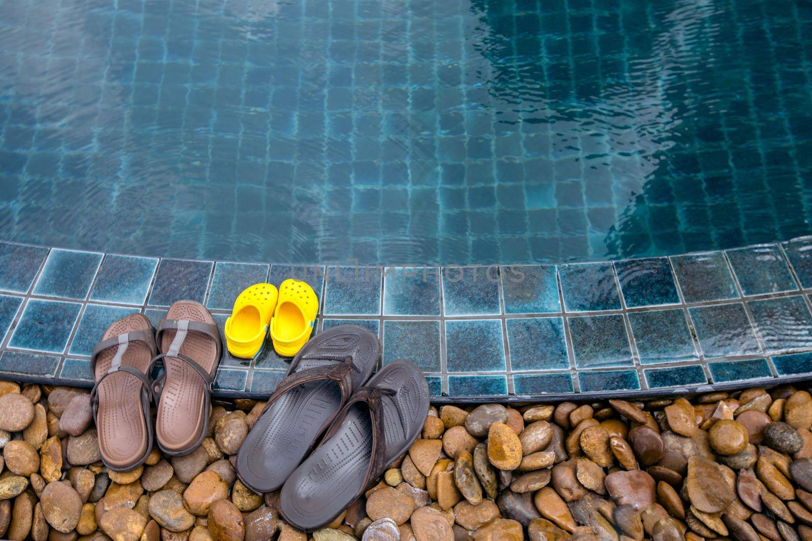 Three pairs of flip-flops of family arranged on wet side of refreshing swimming pool.