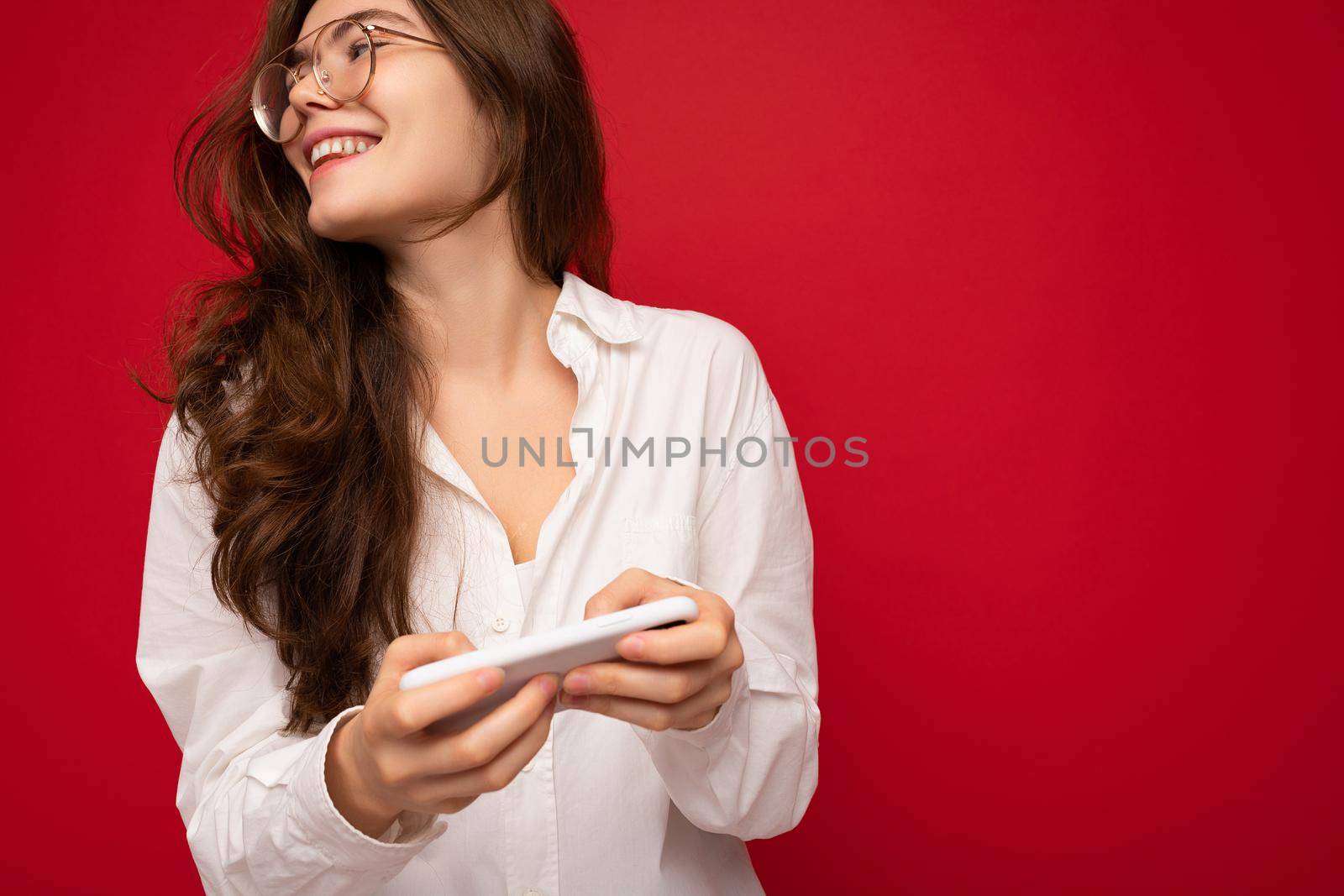 Closeup photo of Pretty smiling positive young brunette woman wearing white shirt and optical glasses isolated over red background holding in hand and using mobile phone playing online games looking to the side.
