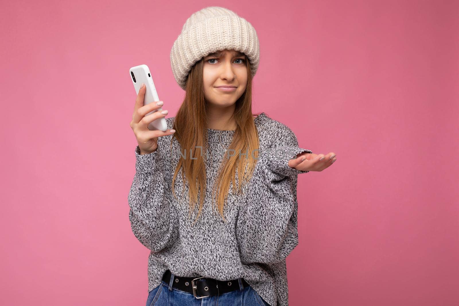 Beautiful funny asking young blonde woman wearing casual grey sweater and beige hat isolated over pink background holding in hand and using mobile phone looking at camera and having doubts.