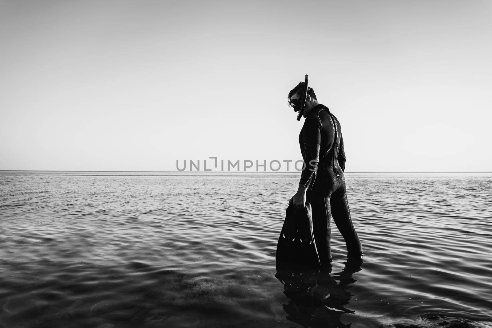 Young man in a diving suit with a mask and flippers goes to the sea in summer outdoor. Monochrome image