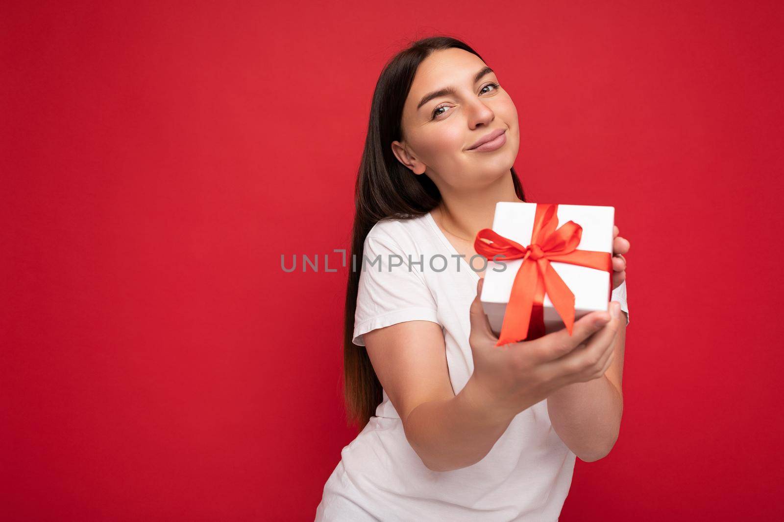 Photo of beautiful positive young brunette woman isolated over red background wall wearing white casual t-shirt for mockup holding white gift box with red ribbon and looking at camera. Free space