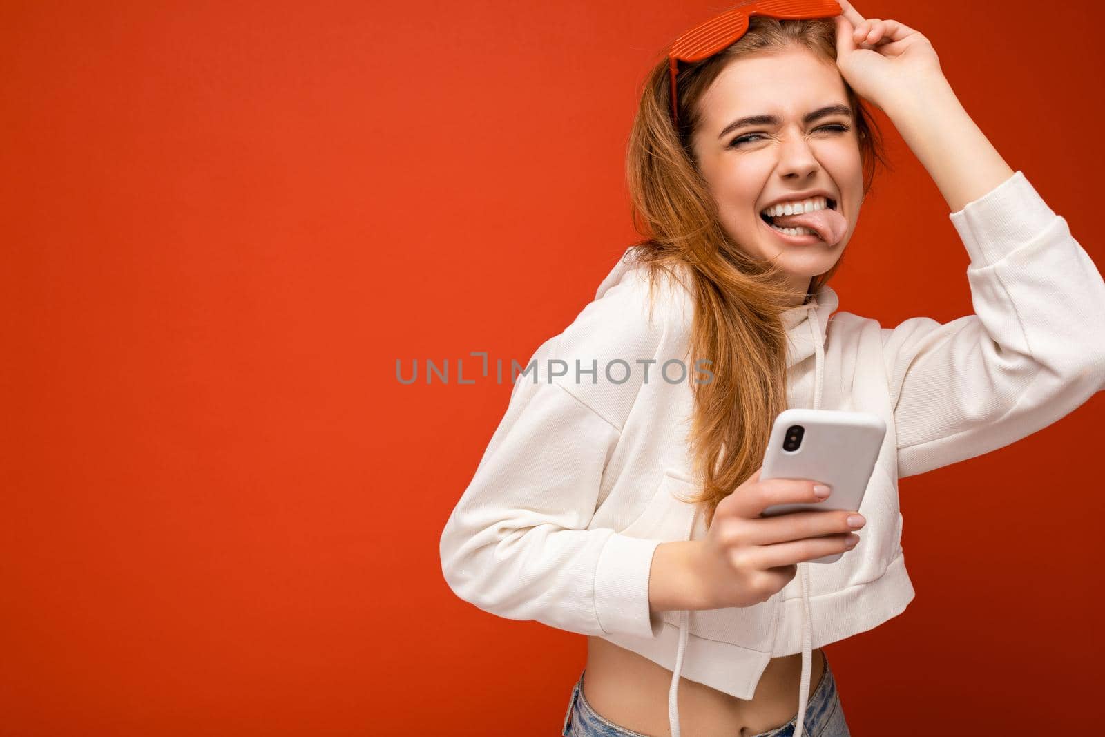 Beautiful emotional young blonde woman wearing stylish white hoodie and funny colorful glasses standing isolated over colourful background holding and using mobile phone communicating online on the internet and showing tongue. Copy space