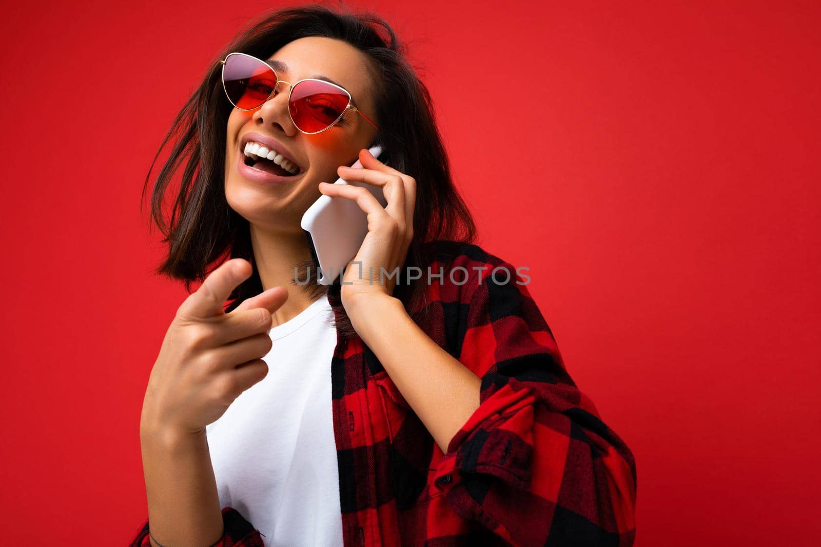 Closeup photo of pretty happy young brunet woman wearing stylish red shirt white t-shirt and red sunglasses isolated over red background communicating on mobile phone looking at camera.