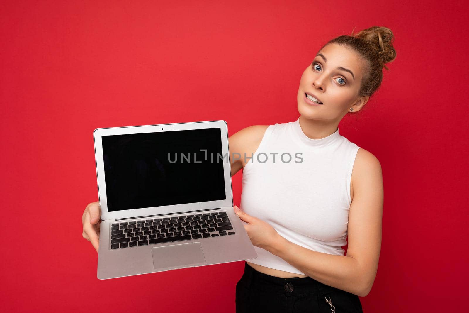 beautiful blond young woman with gathered hair looking at camera holding computer laptop with empty monitor screen with mock up and copy space wearing white t-shirt isolated on red wall background.