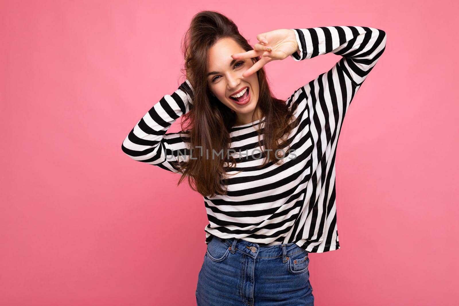 Photo shot of young european positive smiling winsome happy beautiful brunette woman with sincere emotions wearing casual striped pullover isolated on pink background with copy space.