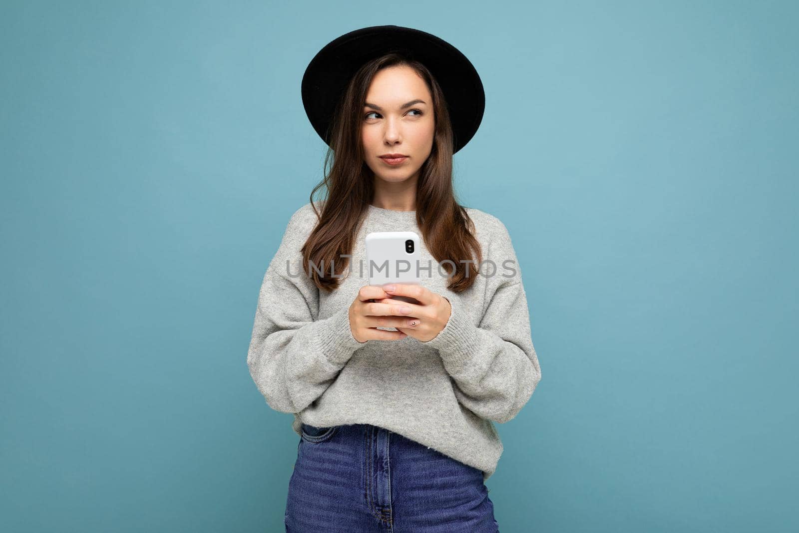 Beautiful young brunette woman thinking wearing black hat and grey sweater holding smartphone looking to the side texting isolated on background.Copy space
