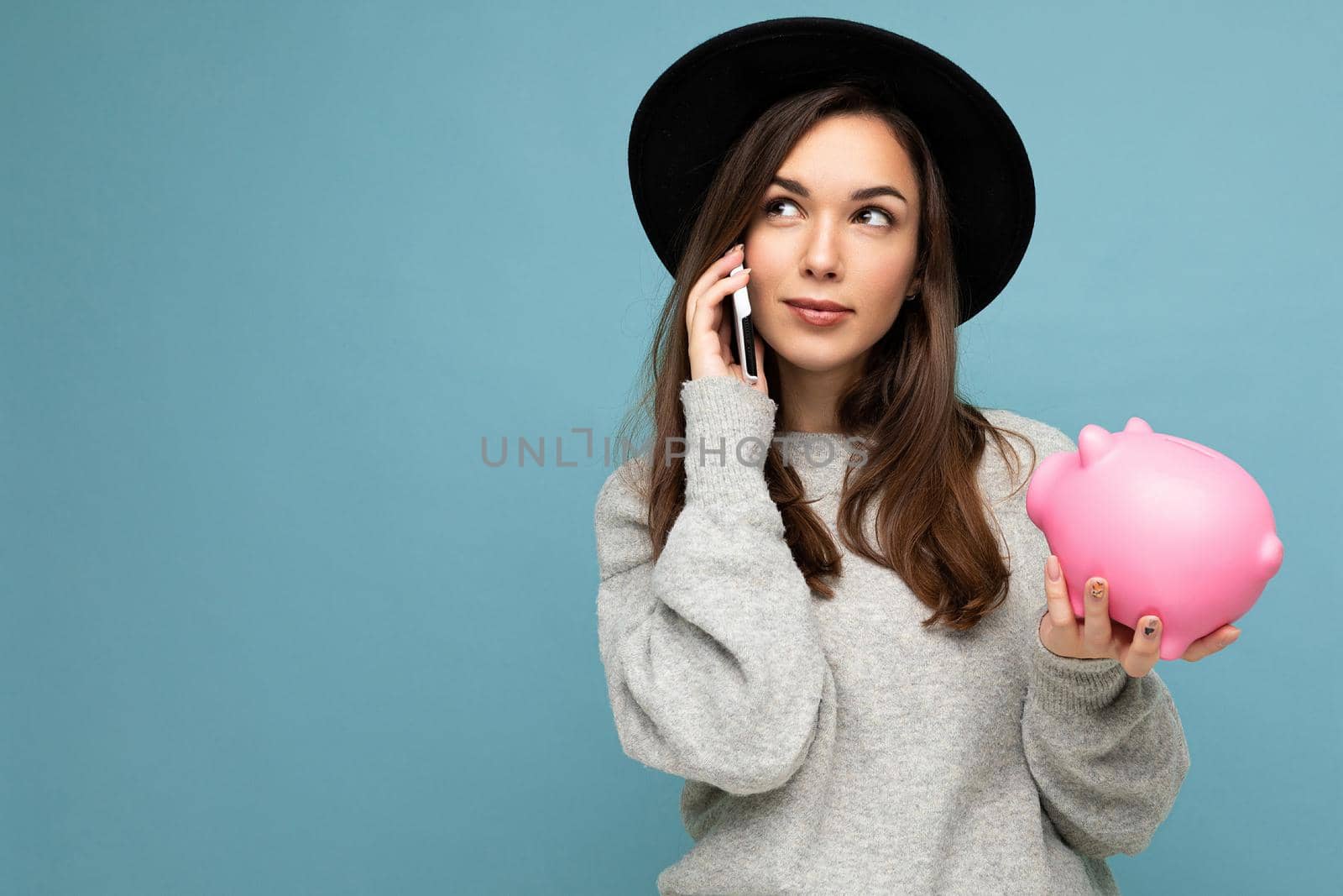 Portrait photo of positive thoughtful dreaming young attractive brunette woman wearing casual grey sweater and black hat isolated over blue background with empty space, holds pink pig penny bank and speaks on mobile phone online.