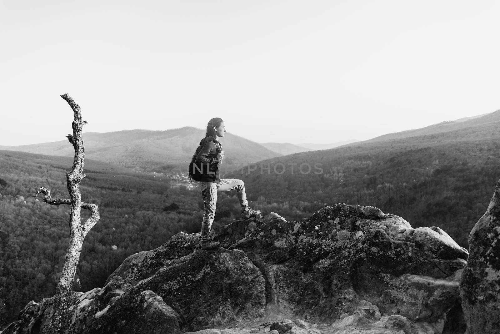 Hiker young woman with backpack standing on peak of rock on background of mountains and looking into the distance in summer outdoor. Monochrome image
