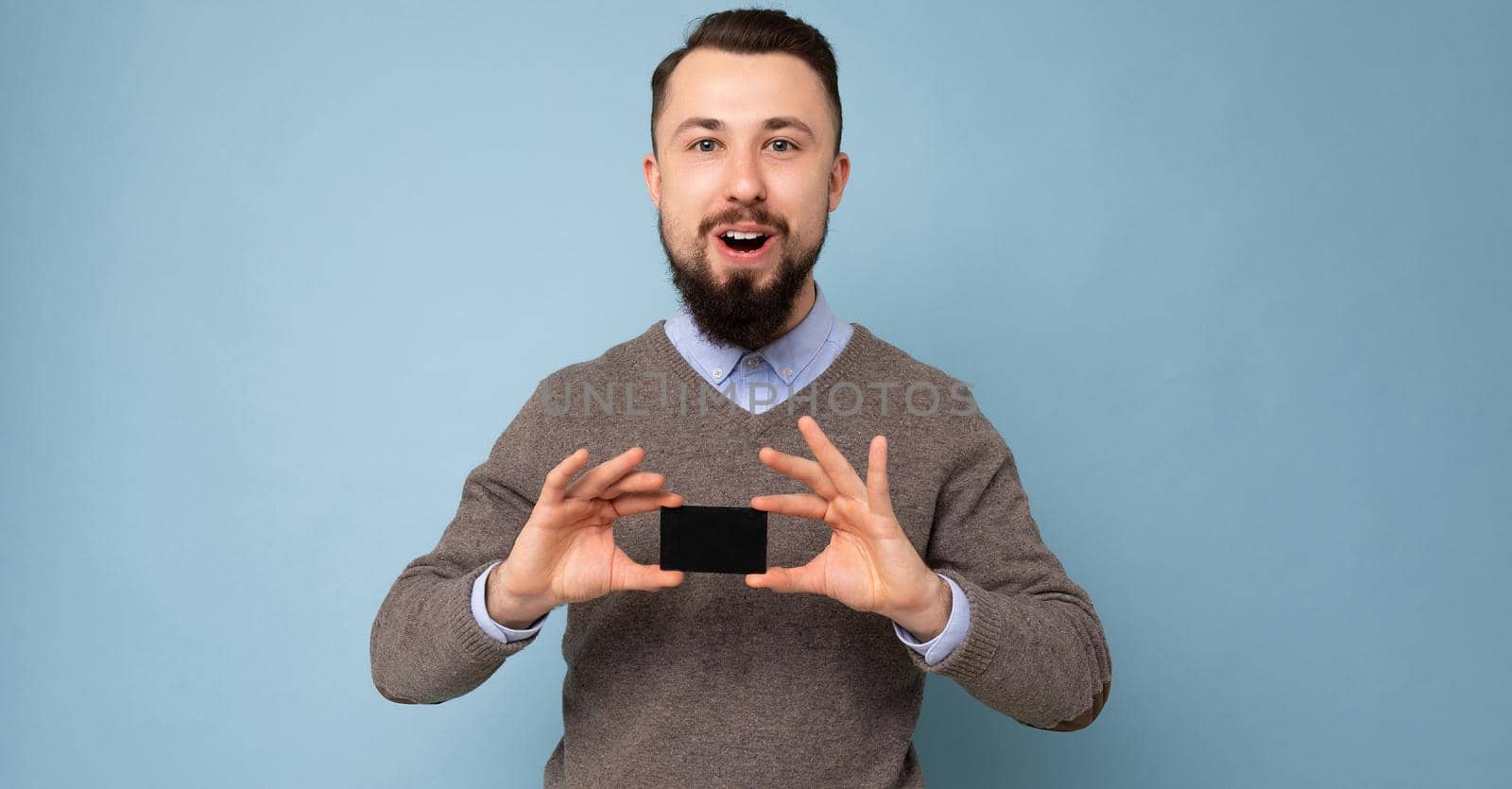 Panoramic photo of Surprised Handsome smiling brunette bearded adult man wearing grey sweater and blue shirt isolated on background wall holding credit card looking at camera.