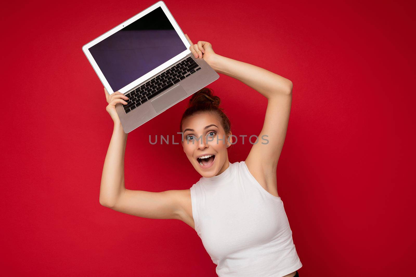 beautiful fascinating happy surorised blond young woman with gathered hair looking at camera holding computer laptop with empty monitor screen with mock up and copy space wearing white t-shirt isolated over red wall background.