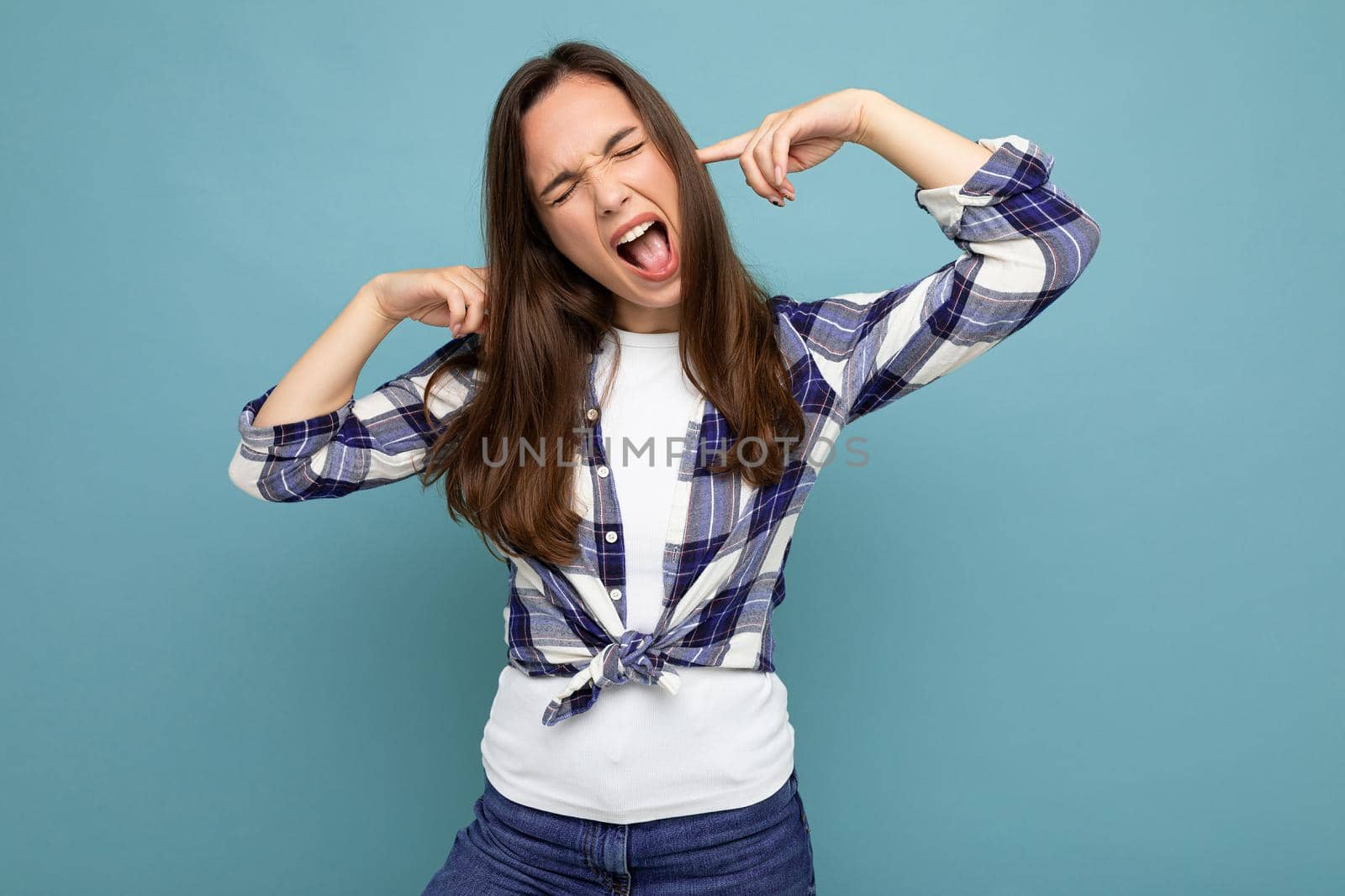 Young emotional attractive brunette female person isolated on blue background with copy space and covering ears and shouting by TRMK