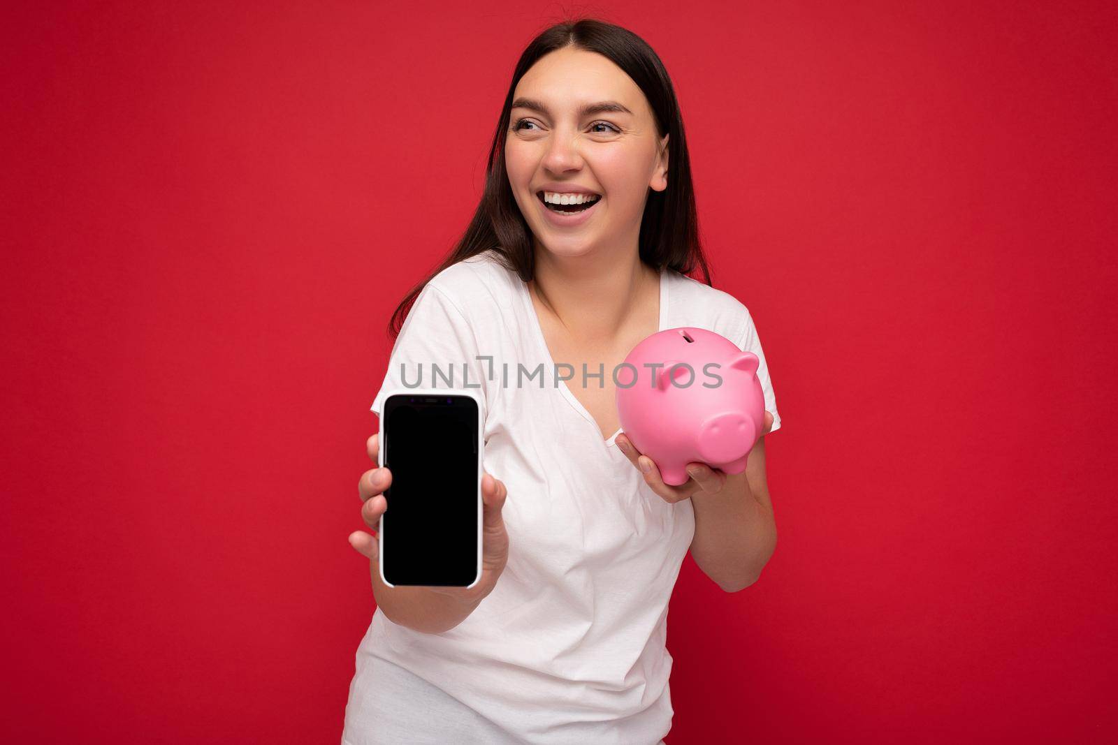 beautiful happy young brunette woman wearing casual white t-shirt isolated over red background with empty space holding in hand mobile phone and showing smartphone with empty screen for mockup and money box looking like pink pig looking to the side.
