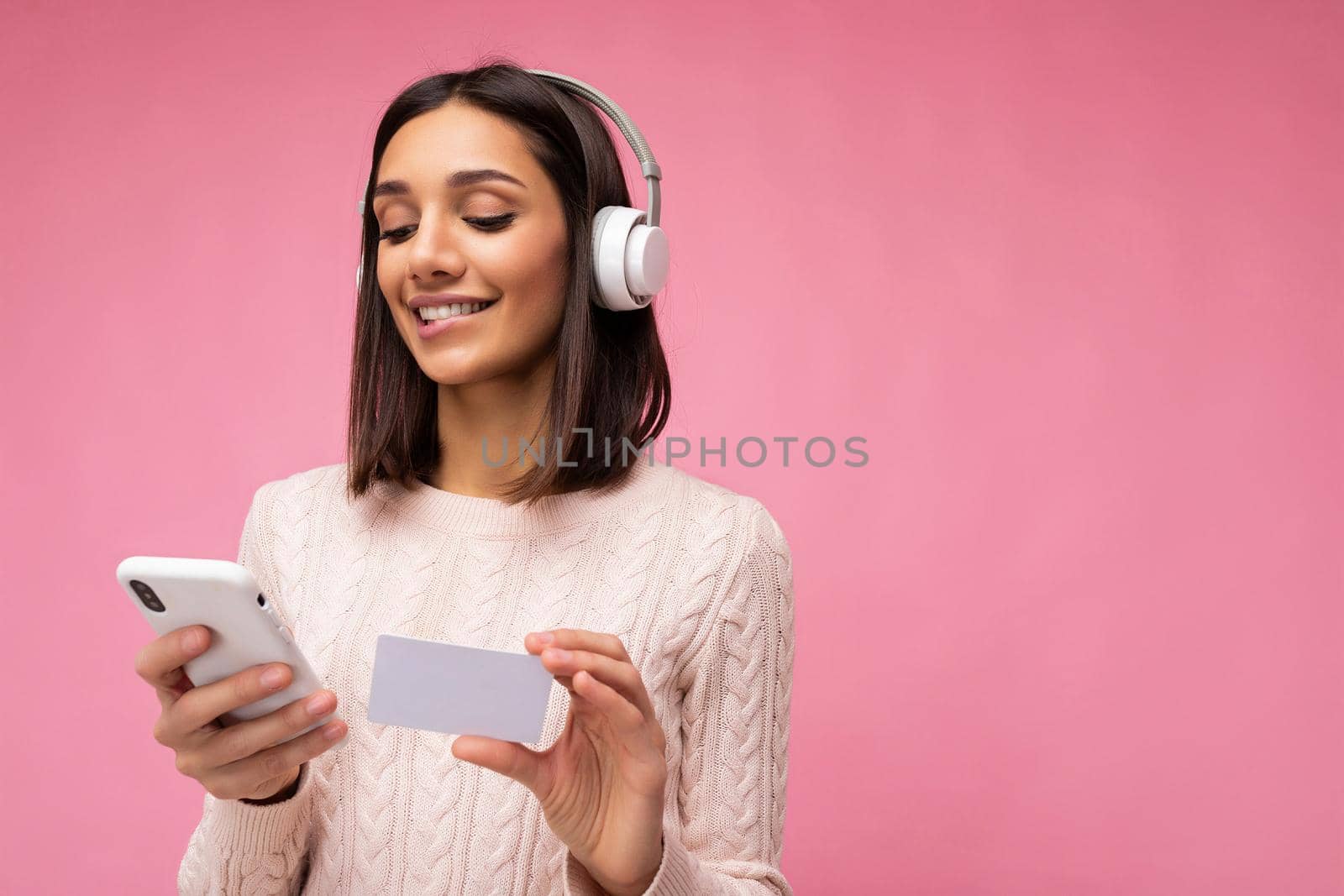 Photo of beautiful positive smiling young brunette woman wearing pink casual sweater isolated over pink background wall wearing white bluetooth wireless headphones and listening to music and using mobile phone making payment online through credit card looking down. copy space