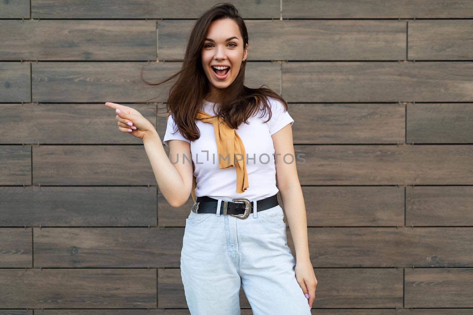 Young emotional positive joyful smiling beautiful brunette woman wearing casual clothes standing in the street near the wall and pointing at empty space. Lifestyle concept.