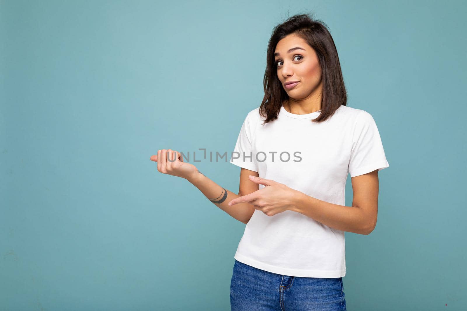 Portrait of young pretty nice winsome brunette woman with sincere emotions wearing casual white t-shirt for mockup isolated over blue background with copy space and pointing at empty space for text.