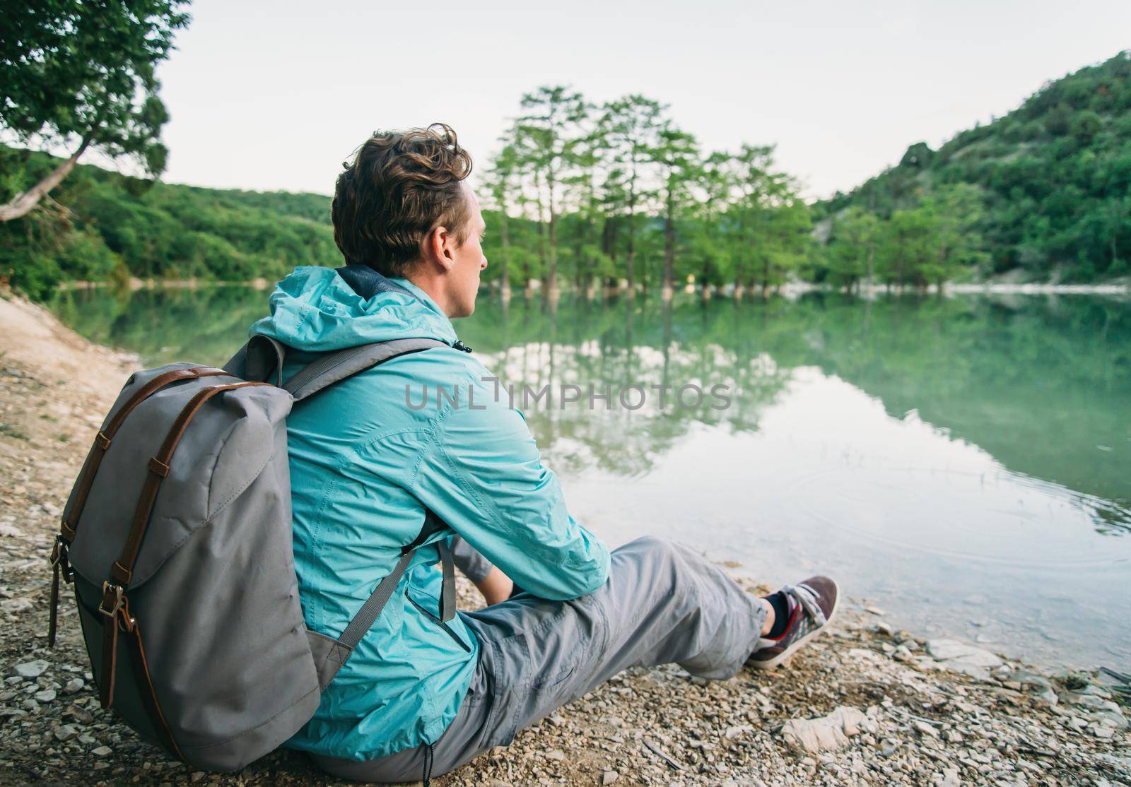 Traveler young man with backpack sitting on shore of lake and enjoying view of nature