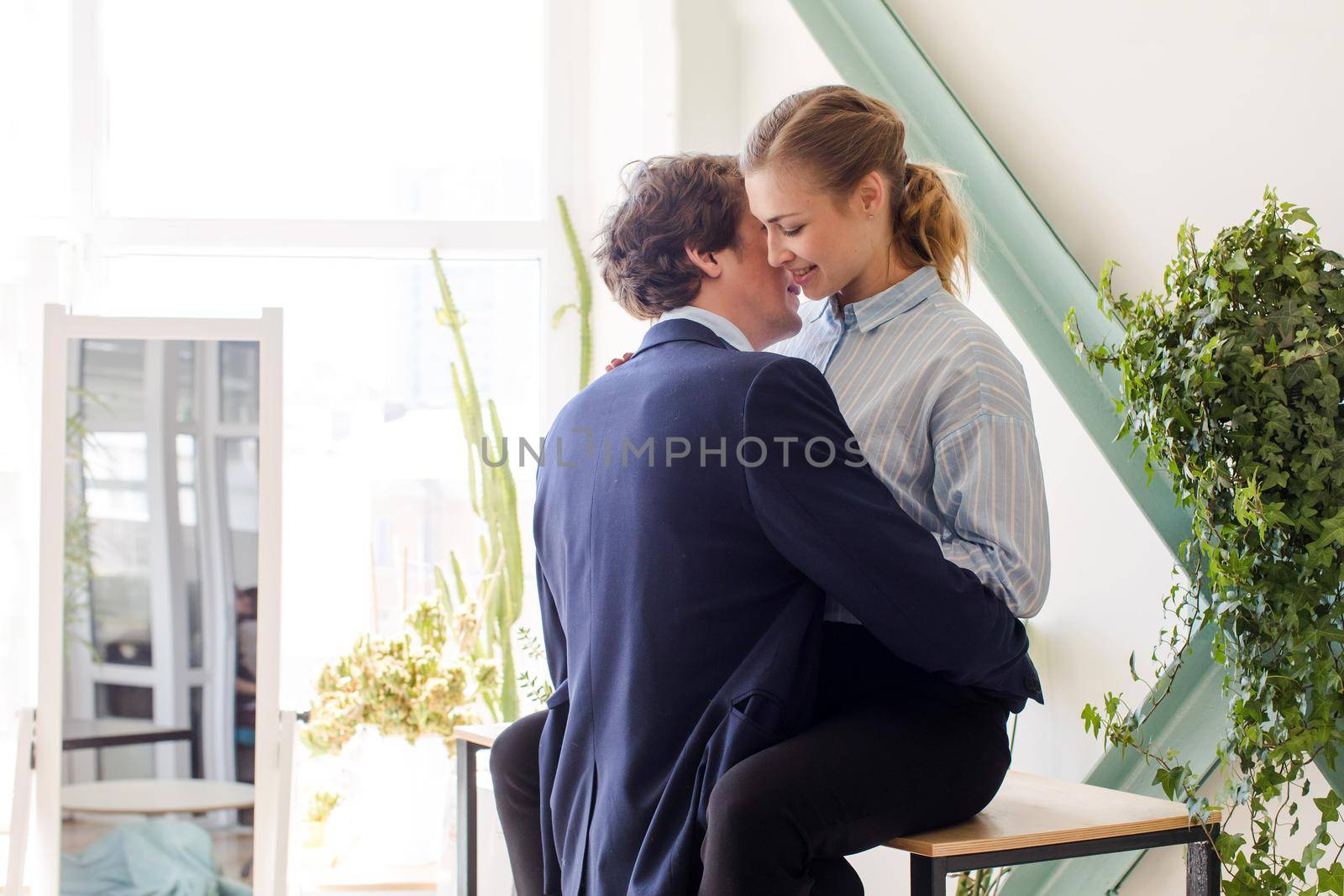 Young man and woman in formal clothes about to have sex in office on table desk among green plants