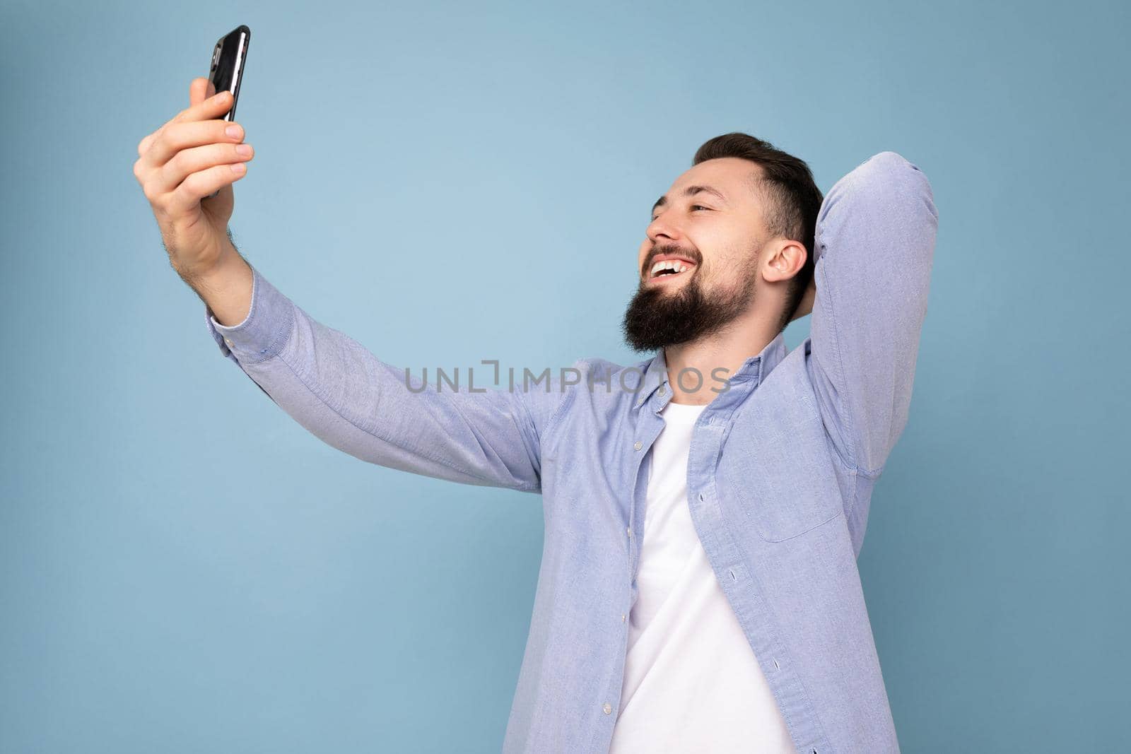 Self-confident handsome young brunet bearded man wearing casual stylish clothes standing isolated over blue background wall holding smartphone taking selfie photo looking at mobile phone screen display.