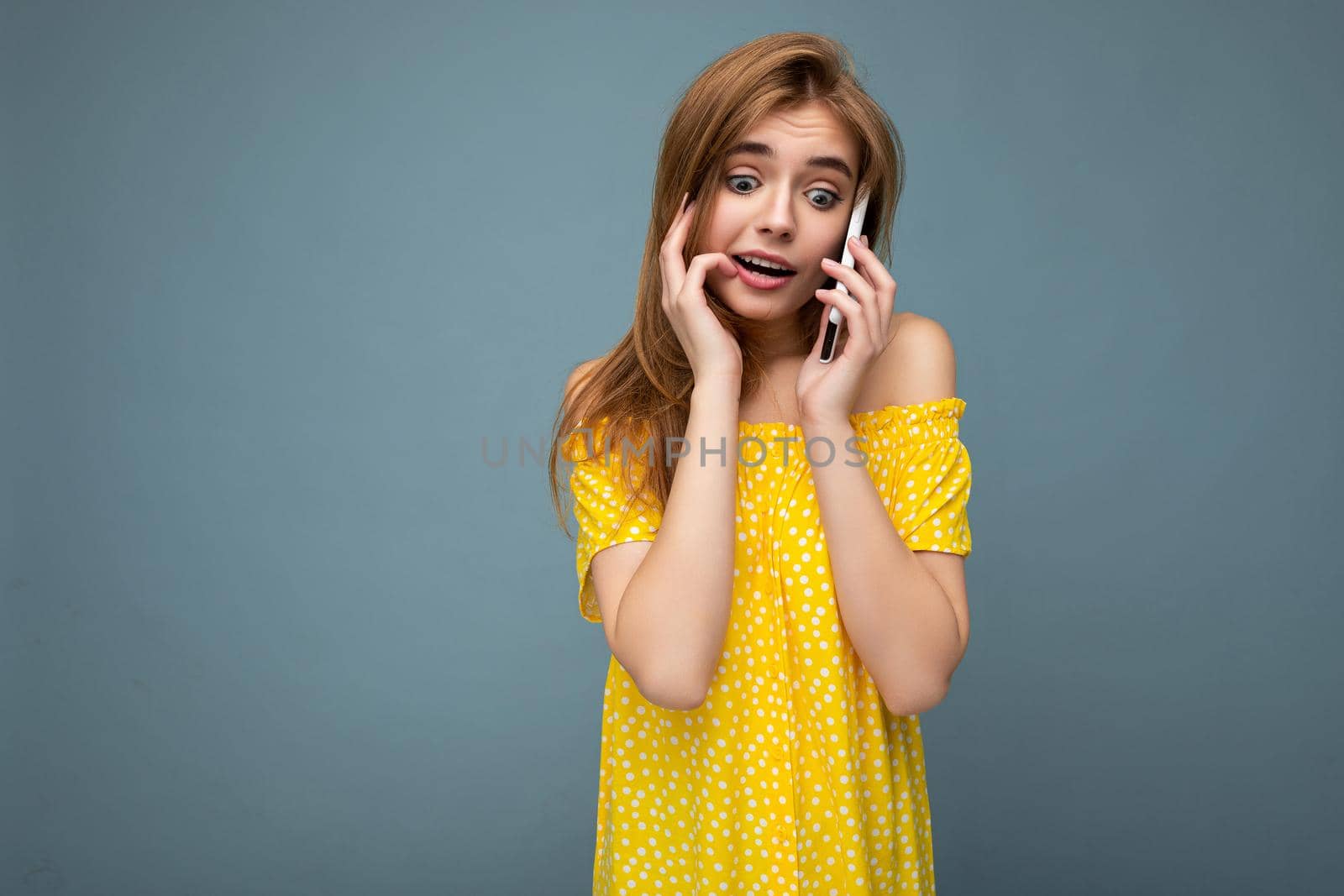 Closeup photo of pretty positive young blonde woman wearing stylish yellow summer dress standing isolated over blue background holding and talking on mobile phone with sincere emotions looking to the side.